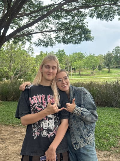 Two individuals are photographed in what appears to be a park or grassy area in Ban Huai Sai, Thailand. The individual in the foreground has long blonde hair, wearing a dark t-shirt with a graphic print and has their eyes closed. They are being hugged from behind by another individual wearing glasses and a denim jacket, who is giving a "thumbs up" gesture with their right hand. The background includes trees, grass, and a cloudy sky.