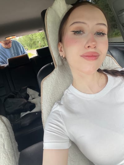 A young woman with light brown hair and makeup is taking a selfie in the backseat of a car. She is wearing a white t-shirt and has a white furry blanket behind her. In the background, a young man wearing a light blue sweatshirt and pink glasses is visible in the front passenger seat. He appears to be looking down at something in his lap. Outside the car window, trees and a road are visible, suggesting a rural or suburban setting. The overall impression is a casual snapshot of a moment during a car ride, possibly in Gavrilovka, Russia, given the provided context.