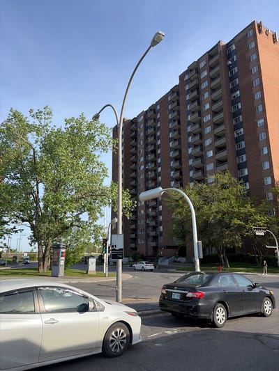 The image captures an urban street scene in bright daylight. A prominent, tall apartment building constructed from brown brick with numerous balconies and windows stands on the left. Several large, leafy green trees are scattered throughout the frame, with one particularly large tree in the foreground and midground.

In the lower right, two cars are visible on the street: a black Toyota Corolla sedan with license plate "YBY XGD" is parked or stopped, and a white Toyota Prius sedan is partially visible in the foreground. The street itself has asphalt pavement and concrete curbs.

The scene features typical street infrastructure, including two prominent streetlights with curved arms, a traffic light displaying a green signal, and various street signs mounted on poles. The sky is clear and blue, and the strong sunlight casts distinct shadows, suggesting it is mid-morning or afternoon. No direct human activity is taking place, although a distant figure is faintly visible behind the black car.

Visible text includes the license plate "YBY XGD" on the black Toyota Corolla. A black street sign above the traffic light displays French text, partially readable as "Lorsque le moteur tourne..." and includes a graphic of a car, indicating it is likely an anti-idling sign.