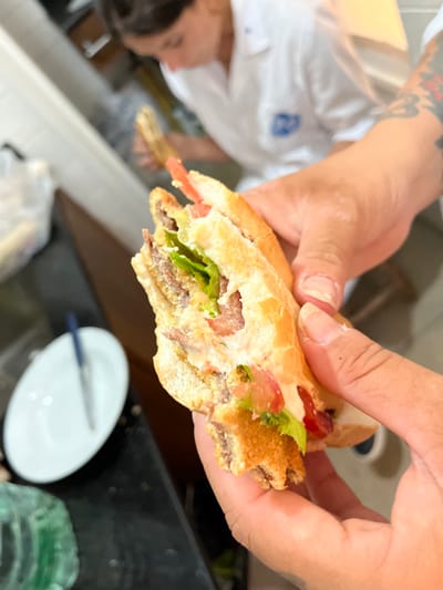 A sandwich is held in two hands. It appears to be a submarine sandwich, cut in half with a portion missing. Lettuce, tomatoes, and meat are visible within the sandwich. A plate with a knife on it is in the background. The hands are fair-skinned with tattooed arms. The background is a kitchen with some cabinets and the person holding the sandwich appears to be wearing a white shirt.