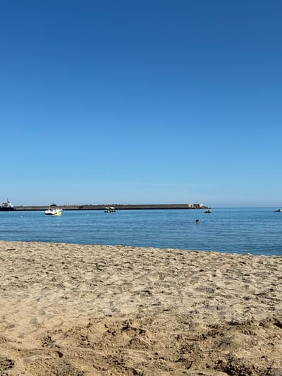 The image shows a beach and the sea on a clear day, likely in the city of Rethymno, Greece, given the context of the provided hints. The foreground is dominated by sand dunes, with intricate patterns of light and shadow. Beyond the sand is the calm, blue sea, stretching towards the horizon. In the distance, a long breakwater or pier extends into the water, with a lighthouse structure visible on its far end. Several boats are scattered across the sea, including a white speedboat with yellow accents, a smaller boat with an awning, and what appear to be jet skis. One person is visible swimming in the water near a jet ski. The sky is a vast expanse of clear, bright blue. The overall scene depicts a peaceful coastal environment.