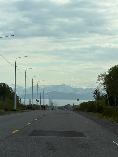 The image depicts an asphalt road with yellow dashed lane markings leading into the distance. The road is lined with numerous lampposts on the left side, and a dense line of trees on the right. In the background, a body of water is visible, beyond which are snow-capped mountains under a cloudy sky. There are no people clearly visible in the scene, though a pedestrian crossing sign is present further down the road on the right. The environment appears to be a semi-urban or rural area outside of the city of Krutoberegovoy, Russia, given the presence of the road, lampposts, and natural landscape. The overall lighting suggests it is daytime, possibly overcast.