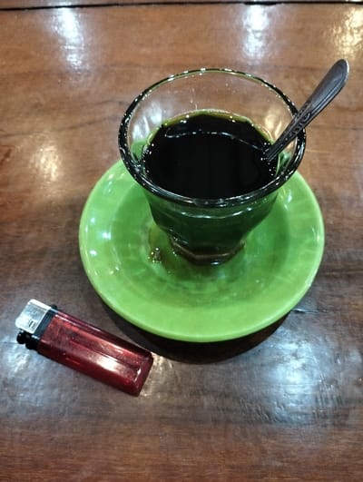 A clear glass containing a dark liquid, consistent with black coffee, is centrally positioned on a bright green ceramic saucer. A silver-colored metal spoon rests vertically within the glass. To the lower left of the saucer, a translucent red plastic disposable lighter with a black top is horizontally situated. All objects are resting on a dark brown wooden table surface, which exhibits a polished, reflective finish and visible wood grain. The scene is located in Jeunieb, Indonesia. No individuals or active interactions are visible.