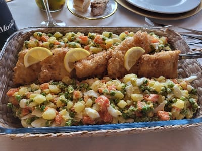 The image displays a close-up of a main course served in a decorative glass dish, nestled within a woven wicker basket or tray. The dish features two primary components: golden-brown, breaded and deep-fried fish pieces, and a colorful potato and vegetable salad.

The fried fish, appearing crisp and golden, is arranged down the center of the serving dish. Several pieces are visible, with two thin, crescent-shaped lemon slices garnishing the fish in the foreground, suggesting a fresh, zesty complement. On the right side, a serving tong is holding one of the fish pieces, indicating the meal is ready to be served or is in the process of being portioned.

Flanking both sides of the fried fish is a generous amount of salad. This salad appears to be a variation of a potato salad, possibly Russian salad (Salad Olivier), characterized by diced yellow potatoes, bright green peas, orange diced carrots, and small pieces of white, firm-looking hard-boiled egg. The ingredients are mixed together, likely with a creamy dressing, though the dressing itself is not distinctly visible.

In the background, elements of a dining setting are visible, slightly out of focus. To the upper left, part of a dark bottle, possibly a wine bottle, can be seen, with the word "Monte" partially visible on its label. Adjacent to the bottle, a stemmed glass holds a yellowish beverage, possibly juice or a cocktail. Further back and to the right, a small dessert plate with a golden rim holds two small, neatly cut pieces of what looks like cake or pastry. A stack of white plates with golden rims and various pieces of silverware (forks and knives) are also visible on a light-colored tablecloth, suggesting a formal or semi-formal dining occasion. The overall scene conveys a pleasant gathering or family meal.