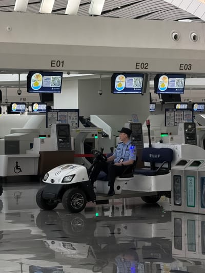 The image depicts an indoor scene, likely an airport, with multiple check-in counters labeled E01, E02, and E03. Several digital screens displaying "Free Wi-Fi" and QR codes are visible above the counters. A police officer in uniform is seated in a small, white electric vehicle, which is positioned between two check-in counters. The vehicle has the word "POLICE" and Chinese characters printed on its side. The floor is highly reflective, showing mirror images of the surrounding environment. In the background, there are security cameras and other airport infrastructure. The overall scene suggests an operational check-in area within an airport in Guangyang District, China.