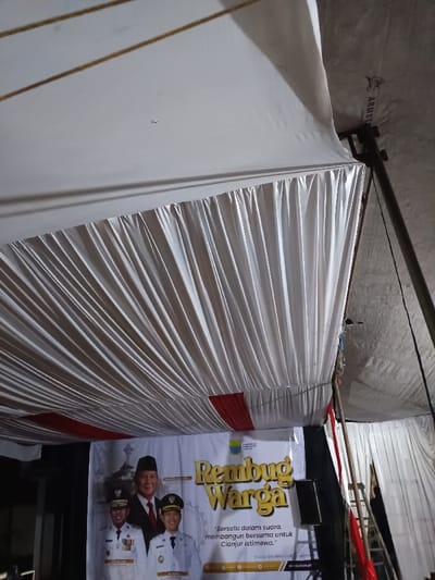 The image displays an interior view of a covered event space in Kawungluwuk, Indonesia, likely a tent. The ceiling is draped with white pleated fabric, creating a textured overhead surface, with segments of red fabric visible on the lower left portion of the drapes. A dark support pole is present on the right, extending upwards, with a rougher, possibly insulated, tent material visible above the decorative white ceiling. In the lower half of the frame, a large white banner serves as a backdrop. It features the gold-colored text "Rembug Warga" (Citizen's Discussion) and the slogan "Bersatu dalam suara, membangun bersama untuk Cianjur Istimewa" (United in voice, building together for a special Cianjur). The banner displays three portraits: a central older man wearing a peci (traditional cap) and a dark suit, flanked by two younger men in white uniforms adorned with epaulets and medals, both smiling. A dark, rectangular speaker or monitor is positioned on a stand in front of the banner on the right side.