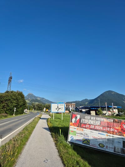 The image depicts a road scene in Niederndorf, Austria, on a clear day with a bright blue sky. In the foreground, a paved road curves to the left, with a sidewalk running alongside it. The sidewalk is bordered by green grass and wildflowers. To the right of the road, a large banner is displayed, advertising the "FEST DER FREIWILLIGEN FEUERWEHR NIEDERNDORF" (Festival of the Volunteer Fire Department Niederndorf) on August 1, 2026, starting at 6:00 PM. The banner also features the logos of various sponsors. Further down the road, a roundabout sign indicates directions to Oberaudorf and Erl (via road 209). A gas station with blue and white signage is visible on the right side of the road, with mountains in the background. A tall electrical pylon stands on the left side of the road amidst green trees and hills. The overall scene suggests a rural or semi-urban environment on the outskirts of Niederndorf. There are no people or animals visible in the image.