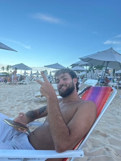 This image captures a beach scene in Cairu, Brazil. In the foreground, a young adult male with dark, curly hair styled in a mullet, a full beard, and a mustache is seated in a multicolored beach chair, directly facing the camera. He is smiling and making a "peace" sign (V-sign) with his right hand. He is shirtless, displaying a tanned complexion with patches of lighter skin on his arms, and a dark tattoo on his left bicep. He is wearing light-colored shorts and holds a gold-colored smartphone in his left hand. The beach chair has a white frame and a seat with horizontal stripes of orange, pink, purple, and blue. The ground beneath him is light-colored sand. In the mid-ground and background, numerous white beach umbrellas are open, shading rows of white lounge chairs, many occupied by other individuals. One female, dressed in a light-colored bikini, is standing with her back to the camera in the mid-ground. Palm trees are visible in the far background. The sky is clear blue with a few faint wisps of clouds, under conditions of natural daylight, possibly late afternoon.