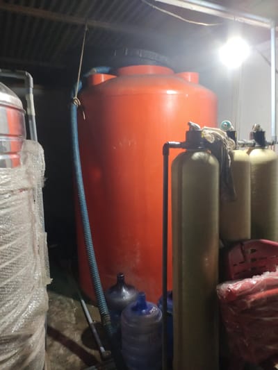 The image shows a cluttered interior space in Teupinkuyun, Indonesia. Dominating the scene is a large orange water storage tank positioned next to water filtration equipment. Several blue water jugs sit near the base of the tank. The lighting is poor and uneven, with a bright light source near the right of the frame. Other objects include a wrapped item on the left and a stacked red item on the right, further contributing to the cluttered environment.