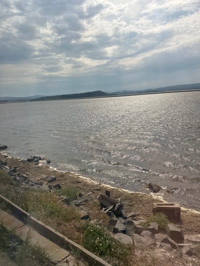 The media file shows a body of water with a rocky shoreline on the right. The water is sparkling due to sunlight reflecting off its surface. The shoreline is covered with dry grass and scattered rocks, with some greenery and small white flowers visible. There are also some concrete structures on the shore.
In the background, there is a distant landmass with hills or mountains, and a hazy horizon. The sky is filled with clouds, some of which are wispy and streaked, suggesting a partly cloudy or overcast day. The overall scene depicts a natural landscape with a body of water.
The image is oriented vertically, with the water occupying the left side of the frame and the shoreline on the right. The lighting suggests it is daytime, likely late morning or afternoon, given the bright sunlight on the water. There are no people or animals visible in the image. No text is present. The location context provided is Port-la-Nouvelle, France, which suggests this could be a coastal area or a large lake. The presence of rocks and the expanse of water could align with such a location.