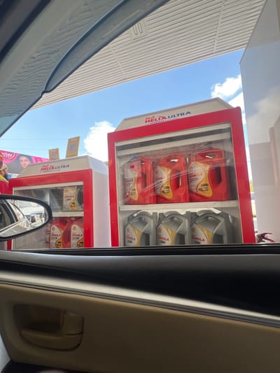The image depicts a scene at a Shell gas station in Abbottabad, Pakistan, viewed from inside a vehicle, specifically from the front passenger side. The foreground shows the beige and dark gray interior door panel of the vehicle, with a beige door handle and an exterior side mirror reflecting a blurred background.
The primary focus is on two prominent Shell lubricant display cabinets positioned outdoors under a gas station canopy. Both cabinets are predominantly red and white, featuring "Shell HELIX ULTRA with PUREPLUS Technology" branding at the top. The right cabinet, fully visible, contains two rows of Shell Rimula lubricant containers behind a clear panel. The upper shelf holds three red containers, while the lower shelf holds three grey containers. A red fire extinguisher is partially visible to the lower right of this cabinet.
The left display cabinet, partially obscured by the vehicle's A-pillar, also contains red and grey Shell Rimula containers. Above this cabinet, two square signs with QR codes are visible.
In the background, a large billboard with a woman's face on a pink background is visible, positioned beyond the gas station structure. To the left of the left display cabinet, a person wearing a yellow and red uniform, likely a gas station attendant, is partially visible from the chest down, facing away from the camera. The sky is bright blue with scattered white clouds, indicating a clear, sunny day. A white, corrugated canopy structure extends overhead, typical of a service station.