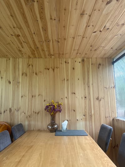 The image depicts an interior scene, likely a dining area, characterized by extensive use of natural wood paneling on both the walls and the ceiling. The wood appears to be light-colored pine, with visible grain patterns and knots. A light-toned wooden table dominates the foreground, and on it rests a glass vase containing a bouquet of dried flowers, predominantly in shades of yellow and purple. To the right of the vase, a small stack of white napkins in a clear glass is visible, placed on a dark gray placemat. Several dark gray upholstered chairs are positioned around the table, with at least two fully visible. In the background, a window with horizontal blinds allows diffused light to enter the room, suggesting it might be daytime. The overall ambiance is rustic and natural. The provided image does not contain any specific landmarks or details that would allow for the identification of the location within Hlevakha, Ukraine.