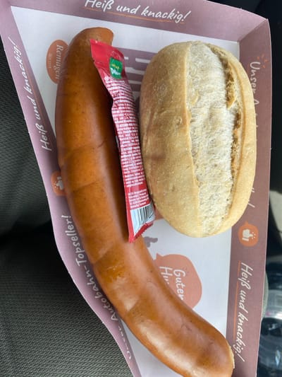 The image displays a meal consisting of a single large, cooked sausage and a bread roll, presented on a branded cardboard tray within what appears to be a vehicle. The primary subject is a reddish-brown sausage, approximately 20-25 cm long, positioned horizontally across the center-left of the frame, exhibiting a slight sheen and subtle grilling marks. Resting atop the sausage, near its upper end, is a red, partially open sachet of Wolf brand ketchup or sauce, displaying white text detailing nutritional information and ingredients in multiple languages, along with a barcode. To the right of the sausage is a single white bread roll (Brötchen), approximately 10-12 cm long, with a light golden crust and a prominent central seam.

These food items are contained within a white cardboard tray featuring brown borders and promotional text. Visible text includes "Heiß und knackig!" (Hot and crunchy!), "Unser Autobahn-Topseller!" (Our Highway Top-seller!), and "Remagen SEIT 1718" on the left border, along with partial text "Guter Hu..." on the lower right interior. The tray is placed on a dark, textured surface, likely a car seat or dashboard. No individuals are present in the scene, and no specific actions or interactions are depicted beyond the static arrangement of the meal. The context suggests a purchase from a highway service station, located in Kammerstein, Germany.