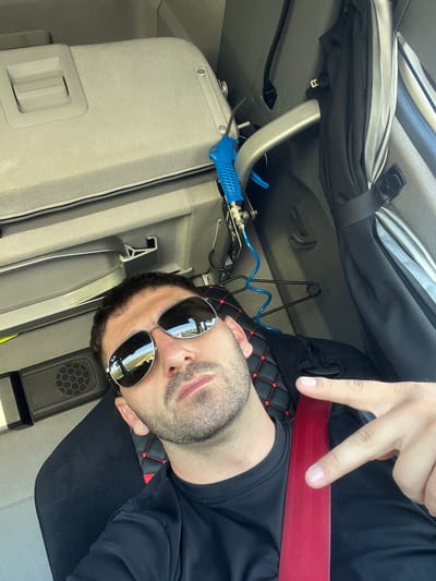 A man is taking a selfie inside a vehicle. He is wearing a black shirt and sunglasses, and has a beard. His right hand is raised, and he is pointing two fingers towards the camera. A red seatbelt is visible across his chest. The interior of the vehicle appears to be a truck cab, with padded seats and overhead storage. A blue air hose and hanger are visible in the background. The reflection in his sunglasses shows a road with some greenery. The image was taken in Mocsa, Hungary.