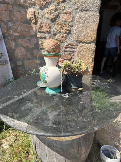 The image depicts an outdoor scene, possibly a patio or garden area, in Santa Cristina d'Aro, Spain. A round, dark, reflective table with a glass top sits on top of a wooden barrel. On the table are decorative objects: a white ceramic vase with a green and red floral pattern, and a black pot containing a succulent plant. A coconut shell is placed on top of the vase. In the background, there is a stone wall with a rough texture and an arched doorway leading into a dark interior. A person wearing a light blue t-shirt and denim shorts is visible walking through the doorway. The ground surrounding the table is covered with dry grass and some small plants. The overall impression is one of a rustic outdoor setting.