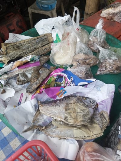 A circular green tray holds a variety of food items, seemingly for sale at a market. Dried fish wrapped in newspaper and other paper scraps are prominent. Several clear plastic bags contain what appears to be dough or flour and pieces of meat or vegetables. A partially visible blue and white checkered surface is at the bottom left and a red plastic basket is in the foreground. In the background, there is a clear plastic container on top of a white stool.