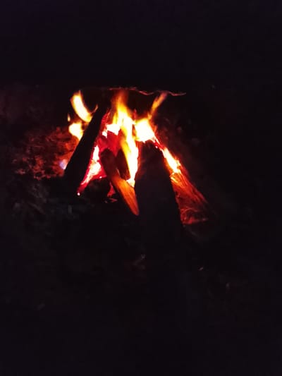 A bonfire burns intensely in the dark, casting a warm, orange glow. Several logs are stacked in a pyramid shape, with flames licking at their edges and rising upwards. The surrounding ground is dark and appears to be covered in dirt and embers. The image is shot at night, and the darkness emphasizes the brightness of the fire. No people are visible in the scene. This scene could be located in Rumuokwuta, Nigeria, a location known for its vibrant community and outdoor activities.