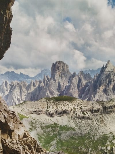 This media file depicts a majestic mountain landscape under a cloudy sky. The scene is dominated by jagged, rocky peaks that are partially covered in snow. In the foreground, a steep, rocky cliff face is visible on the left, adding depth and a sense of scale to the image. The middle ground features a rolling mountain slope with patches of green grass and sparse vegetation, interspersed with rocky outcrops. Further in the distance, more layered mountain ranges stretch across the horizon, their forms softened by atmospheric haze. The sky is filled with a mix of dark and light gray clouds, suggesting a dynamic weather system. The lighting indicates it is likely daytime, with the clouds obscuring direct sunlight, creating a diffused and somewhat dramatic illumination of the landscape. There are no visible people, objects, or text in the image. The overall impression is one of rugged natural beauty and the immense power of geological formations.