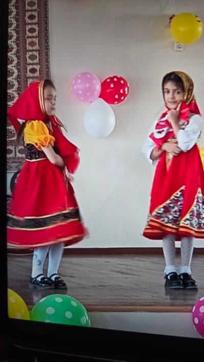 The image shows two young girls dressed in traditional red and gold costumes, possibly performing a dance. The girl on top is wearing a red dress with gold trim and a headscarf, and she is smiling at the camera. The girl on the bottom is wearing a similar costume, but her head is bowed, and she appears to be in motion, with her dress swirling around her.
They are surrounded by balloons of various colors: yellow, pink, red with white hearts, white, and green with white polka dots. A tapestry-like fabric with a geometric pattern is visible in the lower part of the image, suggesting an indoor setting, possibly a cultural event or performance space. The overall mood appears festive and celebratory. The image quality is not very high, with some blurriness and distortion, likely due to it being a photograph of a screen. There is no visible text in the image.