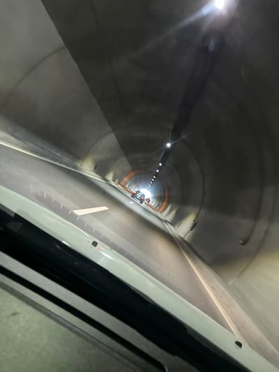 The image depicts the interior of a tunnel, likely a road tunnel, with a perspective from inside a vehicle. The tunnel walls are made of concrete and are illuminated by artificial lights, creating reflections on the road surface. There are lane markings visible on the road. Further down the tunnel, another vehicle can be seen, and at the far end, a brighter light source indicates the tunnel exit. The angle of the shot suggests a wide-angle lens or a fisheye effect, distorting the perspective. The scene is devoid of people other than implied occupants of the vehicles. The environment is sterile and functional, typical of modern tunnel infrastructure. The specific location in Blatino, Bulgaria, cannot be determined from the visual information alone, as there are no identifying landmarks or signs.