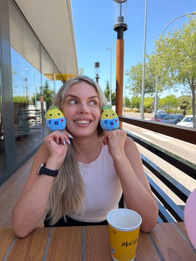 A fair-skinned woman with blonde hair is sitting at a wooden table on an outdoor patio. She is wearing a light pink tank top and black bottoms. On her left wrist, she is wearing a black smart watch with a black strap. She is holding two small, plush pineapple toys up to her head, one on each side. The toys have blue bodies with white bands, and the number "5" is visible on each. The woman is smiling and looking to her right. In front of her, on the table, is a yellow McCafé paper cup. Behind her, there is a railing and trees. The sky is clear and blue. The scene appears to be in a commercial area with a building and lampposts visible in the background, suggesting it's likely a McDonald's with an outdoor seating area.