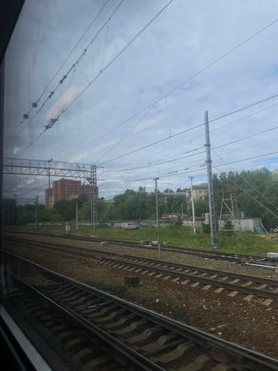 The image shows a view from a train window, looking out at a railway yard and the surrounding environment. In the foreground, multiple sets of train tracks are visible, running parallel to each other and disappearing into the distance. The tracks are laid on ballast and surrounded by grass and weeds. In the middle ground, there are overhead power lines supported by metal structures and poles. Beyond the tracks and power lines, there is a dense line of green trees and bushes. Further back, several buildings are visible, including a multi-story brick apartment building and smaller industrial or utility structures. One building appears to be a light-colored, somewhat dilapidated structure with a metal ladder attached to its side. A white utility van is parked near one of the buildings. The sky is overcast with a mix of clouds, suggesting a cloudy or rainy day. The overall scene depicts a typical view of the urban fringe or an industrial area adjacent to a railway line. The presence of overhead electric lines indicates that this is likely an electrified railway line. The location is identified as Aprelevka, Russia.