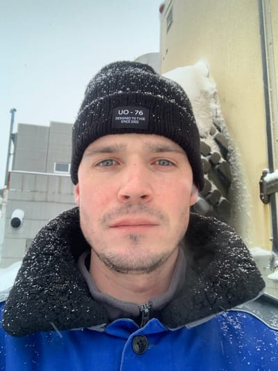 A fair-skinned man with blue eyes and stubble wears a black beanie with a "UO-76" tag. He is wearing a blue jacket with a black fleece-lined collar. Snowflakes are visible on his hat and jacket. The background includes a building with snow on its roof and ground. The overall image appears to be taken outdoors in snowy weather.