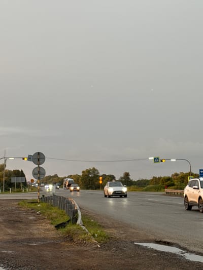 This media file captures a slightly overcast, possibly rainy day in Sovkhoznyi, Russia. The scene depicts a road, likely a highway or a major thoroughfare, with vehicles driving on it. The road surface appears wet, indicating recent or ongoing precipitation.
The primary subjects are several cars, including a white car visible in the upper right portion of the image and another white car with its headlights on in the lower half, suggesting it's either dusk, dawn, or the weather has reduced visibility. Other vehicles are present further down the road, some appearing as headlights. There are traffic lights and road signs visible, including a pedestrian crossing sign and circular traffic signs. Trees and vegetation line the side of the road, creating a natural backdrop. The sky is a muted grey, contributing to the subdued lighting conditions.
The activity taking place is vehicular traffic on a road. The wet road and overcast sky suggest a potentially damp or gloomy atmosphere. There are no people clearly visible in the frame, focusing the attention on the vehicles and the road environment. The presence of traffic signals and road signs indicates an urban or semi-urban setting where traffic management is in place. The overall impression is a mundane scene of travel on a road under less-than-ideal weather conditions.