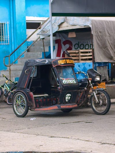 A side-view, full-shot image captures a black and orange tricycle, a common mode of transportation in Cavite City, Philippines, parked on a concrete surface. The tricycle is adorned with several decals, including one that reads "FRUIT DELIVER" in yellow lettering across its front canopy, and another featuring a portrait of Che Guevara. A smaller, stylized white decal appears to read "Jims."

The tricycle is positioned in front of a blue building with a white staircase and railing. Behind the tricycle, a partially visible sign with red and green lettering reads "12 COUNCIL dood". In the background, a wooden crate structure is visible, along with a gray tarp. To the left of the tricycle, a bicycle with a yellow frame and green accents is also parked. The scene appears to be outdoors, likely on a street or in a marketplace, characteristic of the urban landscape of Cavite City. The lighting suggests it is daytime, possibly overcast.