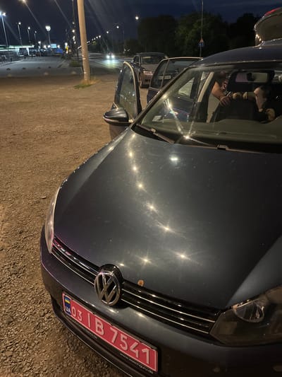 The image shows the front of a dark-colored Volkswagen parked on a gravel lot at night. The car's headlights are on, illuminating the scene. A license plate on the front of the car reads "UA 003 IB 7541". The interior of the car is visible through the open driver's side door, where a person appears to be sitting in the driver's seat, looking at their phone. The background consists of trees and streetlights, suggesting an outdoor setting in an urban or suburban area. The weather appears to be clear, and the time of day is nighttime. There are some reflections on the car's hood, likely from nearby lights. Another vehicle is parked behind the Volkswagen, partially visible. The overall mood is casual and relaxed, with the person in the car engaged with their phone.