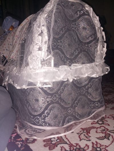 The image depicts what appears to be a baby's bassinet or crib, draped with a patterned fabric and white lace or tulle. The patterns on the fabric appear to be ornate and possibly reflective. The bassinet is on a rug with a floral pattern in shades of brown, white, and red. In the background, to the left of the bassinet, is what appears to be a baby doll or a bundled infant. The scene is indoors, and the lighting is somewhat dim. No specific details confirm the location as Galaosiyo, Uzbekistan.
