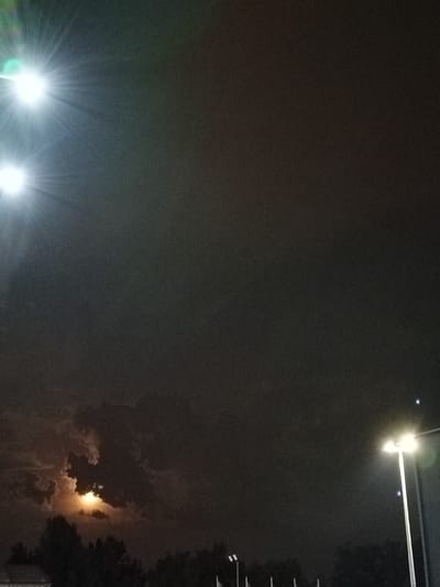 The image captures a nighttime scene in the city of Bytom, Poland. Two bright stadium lights are visible in the upper left corner, casting strong beams. In the lower portion of the image, a partially obscured moon shines through dark, cloudy skies, casting a soft glow on the silhouettes of trees below. A modern streetlamp with two lights stands on the right side, illuminating the surrounding darkness. The overall atmosphere is dark and atmospheric, suggesting an outdoor urban setting at night.