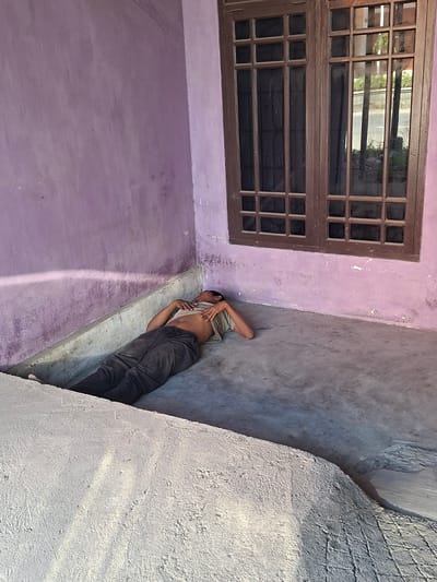 In a corner of a room with purple walls and a concrete floor in Bireuenmeunasahcapa, Indonesia, a person is lying on their back with their eyes closed, appearing to be asleep or resting. The individual is wearing dark pants and a light-colored t-shirt, which is partially pulled up to reveal their abdomen. A window with brown wooden frames and multiple glass panes is visible on the right side of the image. Sunlight streams into the room, casting shadows and highlighting the textures of the walls and floor. The environment suggests an outdoor covered area or a room with open access to the outside.
