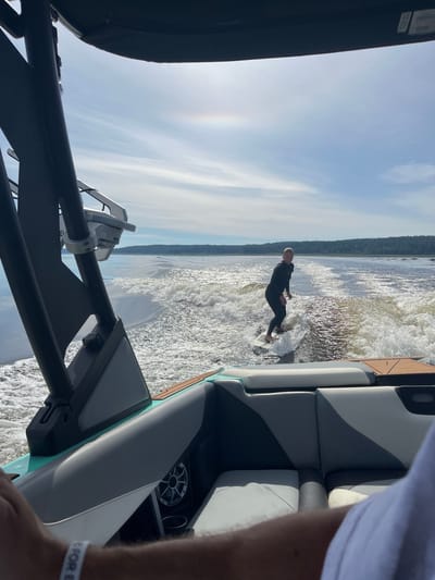 In Gromovo, Russia, a person is wake surfing on a lake, towed by a boat from which the photo is taken. The surfer, wearing a black wetsuit, is positioned on a white board, riding the wake. The boat, partially visible in the foreground, features gray and white seating and a black tower. The water is choppy, reflecting the bright, partly cloudy sky. The background shows a tree-lined shore.