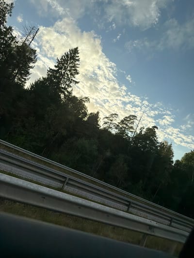 The image captures a view from a vehicle traveling on a road, likely a highway, based on the presence of a metal guardrail in the foreground. The guardrail is silver and shows signs of some weathering and moss growth. Beyond the guardrail, a dense forest with dark green foliage lines the side of the road, suggesting a rural or natural setting. The trees are tall and appear to be predominantly coniferous. The sky above is a mix of blue and white clouds, with some areas of brighter light suggesting the sun is behind or breaking through the clouds. The perspective suggests the photo was taken at an upward angle, looking towards the sky and the tree line. The overall impression is one of a journey through a wooded area on a cloudy day. The location is specified as Oskarsberg, Sweden.