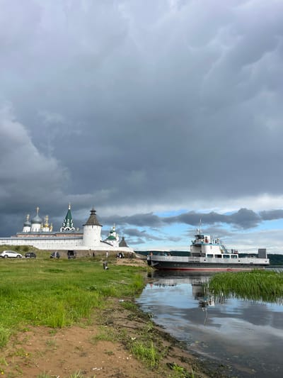 The image depicts a wide river with a ferryboat docked near the shore on the right side. On the left bank, a large, white fortress-like structure with multiple green and gold-domed buildings stands on a hill. Several cars are parked on a dirt road in front of the fortress. The sky is overcast with dark, dramatic clouds, though patches of blue sky are visible. The foreground shows a sandy riverbank with lush green grass growing along the water's edge. The reflection of the ferryboat and the sky is visible on the calm surface of the river. The location is identified as Makar'evo, Russia.