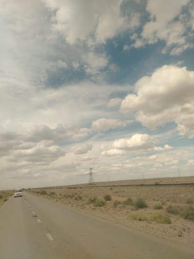 An overcast sky with scattered white clouds is visible over a vast, arid landscape. A two-lane asphalt road stretches into the distance, with a white vehicle driving on the left lane. The roadside is characterized by dry, sparse vegetation and dirt. In the background, a tall lattice tower, likely a power pylon, stands on the right side, and a railway line runs parallel to the road. The scene depicts a journey through a remote, possibly desert or steppe region.