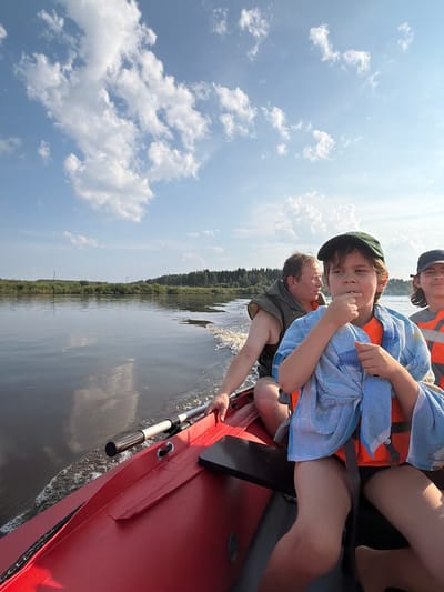 In the city of Kalino, Russia, a red boat glides across a calm river. The boat carries three people. In the foreground, a young boy with a green cap on his head and an orange life vest is wrapped in a blue towel. Next to him, a man in an olive-green life vest is at the helm, steering the boat. Behind them, another person in an orange life vest is partially visible. The riverbank is lined with lush green trees and shrubs, under a bright blue sky dotted with fluffy white clouds. The water reflects the sky and the trees, creating a serene and picturesque scene.