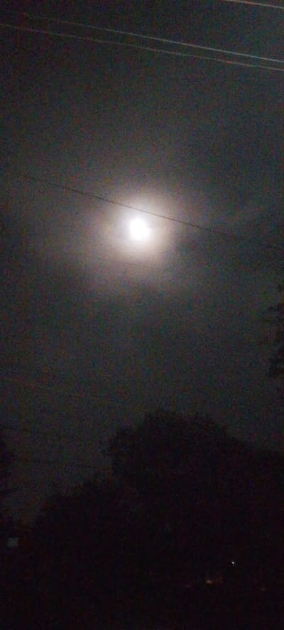 The image depicts a night scene in the Ndenderu ward, Kenya. The sky is dark, and a full moon is visible through a hazy layer of clouds. Electrical wires stretch across the upper part of the frame. Silhouetted trees are present in the lower portion of the image. The foreground is dark, obscuring details on the ground.