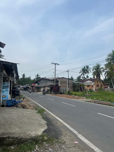 This image depicts a roadside scene in Blanggunci, Indonesia, likely during daylight hours under an overcast sky. The perspective is from the side of a paved road that runs along a row of buildings and vegetation.
The road itself has a white line marking its edge. To the left of the road, there are various structures, including what appear to be small shops or houses with corrugated metal and tile roofs. Some of these buildings have open fronts, revealing merchandise or activities within. Several motorcycles are parked along the roadside in front of these establishments. A black car is also visible further down the road.
Behind the buildings, palm trees and other lush greenery are abundant, suggesting a tropical or subtropical environment. In the background, a larger building with a yellow facade and a red roof is visible, bearing text that reads "IMPONG BLANG GUNCI" and "DESA BLANG GUNCI." This building likely serves as a community center or administrative office.
Overhead, power lines and utility poles crisscross the sky, indicating the presence of electricity infrastructure. The ground beside the road is a mix of concrete, gravel, and grass, with some scattered debris. The overall impression is of a modest, lived-in village or small town setting in Indonesia, with daily life unfolding along the main thoroughfare. There are no prominent individuals in the frame, but the parked vehicles and open shops suggest ongoing activity.