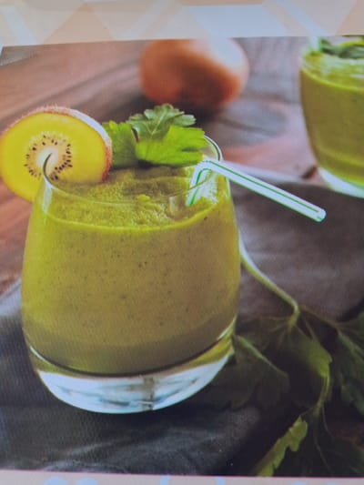 A close-up shot shows a glass filled with a green smoothie, garnished with a slice of kiwi and a sprig of cilantro. A green and white striped straw is inserted into the smoothie. The glass is set on a dark cloth, and in the background, out of focus, are two whole kiwis and some greenery. The image is brightly lit and appears to be a food photograph. There is no indication of Beşik-Jon, Kyrgyzstan in the image.