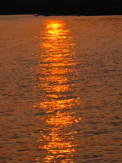 The image shows a body of water reflecting the warm, golden light of a sunset. The surface of the water is rippled, creating a shimmering effect as the light bounces off the crests and troughs. In the distance, a dark silhouette suggests a shoreline or landmass. The overall impression is one of tranquility and natural beauty. The location in the city of Потапово, Russia, is not explicitly visible in the image, but the scene is consistent with a natural setting like a lake or river.