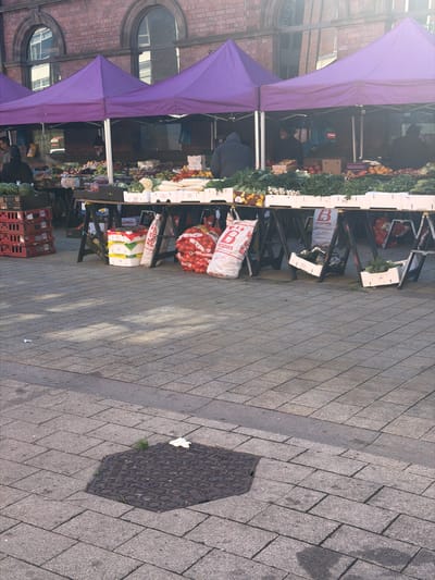 The image displays an outdoor market scene in a paved square in Rotherham, United Kingdom, directly in front of a brick building identified by partially visible text as the "OLD TOWN HALL." Three market stalls with purple canopies are arranged in a line, each supported by wooden trestle tables laden with assorted fresh produce including watermelons, cauliflower, leafy greens, and various fruits and vegetables. Several white polystyrene boxes hold produce on the tables. Underneath the stalls, multiple large white sacks marked "B POTATOES" are visible, alongside stacks of red plastic crates and additional produce containers. Three individuals are discernible behind the stalls: one on the far left, one in the center wearing a dark hooded garment with their back to the camera, and one partially visible on the far right. The ground is paved with light grey rectangular pavers. In the foreground, a hexagonal metal manhole cover is embedded in the paving, with a small piece of white litter nearby. Sunlight casts diagonal shadows across the paved surface from the right.