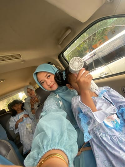 An image captured inside a vehicle in Ketegan, Indonesia, depicts four females. In the immediate foreground, a woman wearing a light blue hijab and a matching long-sleeved dress with pleated cuffs and embroidered white bird motifs looks directly at the camera with a slight smile. Her right wrist, adorned with gold-colored bracelets, is visible at the bottom left. Adjacent to her on the right, a young girl with dark curly hair, wearing a light blue and white patterned dress featuring a character graphic (possibly Elsa from Frozen), holds a small white portable fan to her face, obscuring the upper part of her face. A light blue flower-shaped hair clip is visible in her hair.
Behind these two individuals, another young girl with dark hair tied back, dressed in a similar light blue and white patterned garment, gazes towards the camera. To her right, a second woman, also wearing a pale blue or greyish hijab and matching attire, is seen looking down at a smartphone held in her right hand.
The vehicle's interior features a light-colored headliner with an overhead air conditioning vent and grab handles. Through the vehicle's windows, exterior green foliage, reflections of another vehicle, and sections of buildings are visible under daylight conditions.