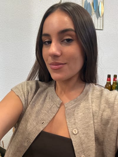 A female individual is depicted in a close-up, chest-up frontal portrait, presenting with long dark hair and brown eyes, and a slight smile. She wears a light beige, short-sleeved button-front cardigan over a dark brown strapless top. The background consists of a textured light grey wall. To the subject's upper right, a metallic wall decoration featuring multiple blue and gold/bronze leaf-shaped elements is mounted. Partially visible on the far right, three bottles are present, consistent with Spanish beer bottles; one is identifiable as Mahou Cinco Estrellas. The scene is indoors, located in Plasencia, Spain.