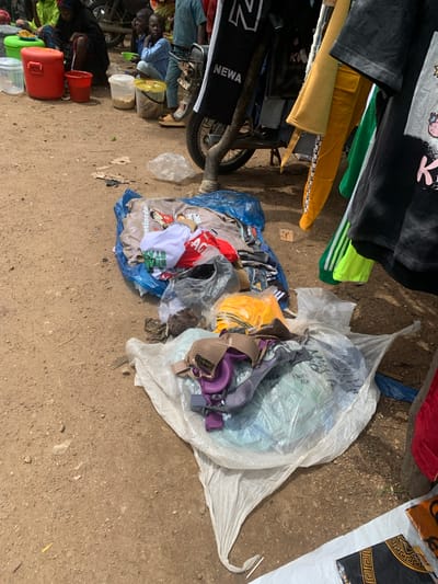 This is an outdoor scene in what appears to be a market in Project, Nigeria. The ground is dirt and appears dusty. In the foreground and midground, there are piles of clothing and other items. To the left, there are several large red and green plastic containers. Behind these containers, several people are seated on the ground. In the center and right of the image, there are items of clothing hanging from a rack or structure, including t-shirts and pants in various colors. Some of the clothing items are also piled on the ground, some wrapped in plastic. A motorcycle is visible behind the hanging clothes. The overall impression is of a busy, informal market setting.