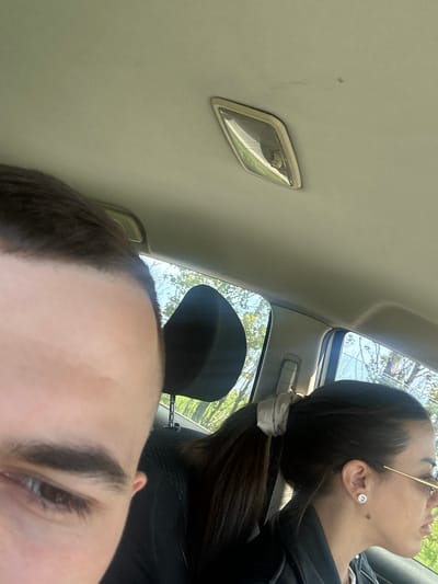 The image captures a casual, close-up perspective from inside a car, likely in Voysil, Bulgaria, during bright daylight. The scene is dominated by two individuals. In the foreground, the top of a person's head, including their forehead, part of an eyebrow, and one eye, is visible, suggesting they are either taking the photo or positioned very close to the camera. Their dark hair is visible at the hairline.

In the mid-ground, another individual is seen in profile, looking upwards and slightly to the right of the frame. This person has dark hair tied back with a light-colored scrunchie. They are wearing gold-rimmed glasses and a prominent diamond stud earring in their ear. A small mole is visible on their cheek.

The background reveals parts of the car's interior ceiling, including a light fixture and a sunroof or side window through which bright blue sky and green tree branches are visible, indicating a clear, sunny day. A dark headrest is also partially visible behind the person in the foreground. The overall impression is one of a relaxed moment within a vehicle, possibly enjoying a journey or parked in a scenic spot. No text is visible in the image.