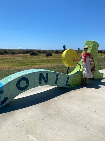 A woman stands next to a large, colorful, ceramic sculpture that spells out "Conil" in the city of Conil de la Frontera, Spain. The sculpture is green and blue, with a yellow circular element attached to it. The woman is wearing a light-colored dress, a red scarf, sunglasses, and a straw hat in her hand, along with a straw bag. She is smiling and looking at the camera. The sculpture is located outdoors on a paved surface, with grass and scrubby vegetation in the background. The sky is clear and blue. In the distance, there are rows of thatched umbrellas, suggesting a beach is nearby.