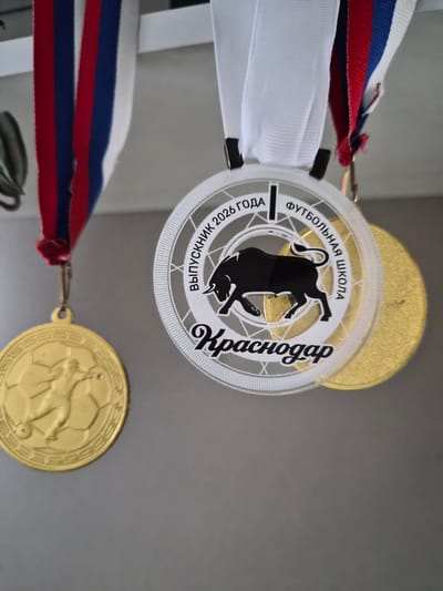 The image shows a close-up of three medals hanging on a gray wall. The leftmost medal is gold and features a relief of a soccer player in action, with a soccer ball pattern around its edge. It is hanging from a ribbon with the colors of the Russian flag (white, blue, and red). To its right is a round, clear medal with a black silhouette of a bull and text that reads "ВЫПУСКНИК 2026 ГОДА" (GRADUATE 2026) and "ФУТБОЛЬНАЯ ШКОЛА Краснодар" (FOOTBALL SCHOOL KRASNODAR). This medal is also hanging from a white ribbon. Behind the clear medal, part of another gold medal is visible. The overall scene appears to be indoors, with the medals likely being awards or mementos. There are no people or specific city locations of Lenina, Russia explicitly depicted.