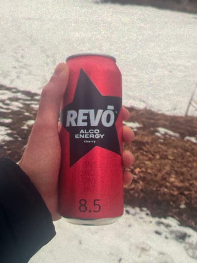 A person's hand is holding a red can of Revo Alco Energy Cherry. The can has a black star logo with the word "REVO" in white letters. Below that, it says "ALCO ENERGY" and then "cherry" in smaller letters. The number "8.5" is printed in black at the bottom of the can. The background appears to be a snow-covered area with brown patches of ground visible. The image is grainy. The location is Paczkowo, Poland.